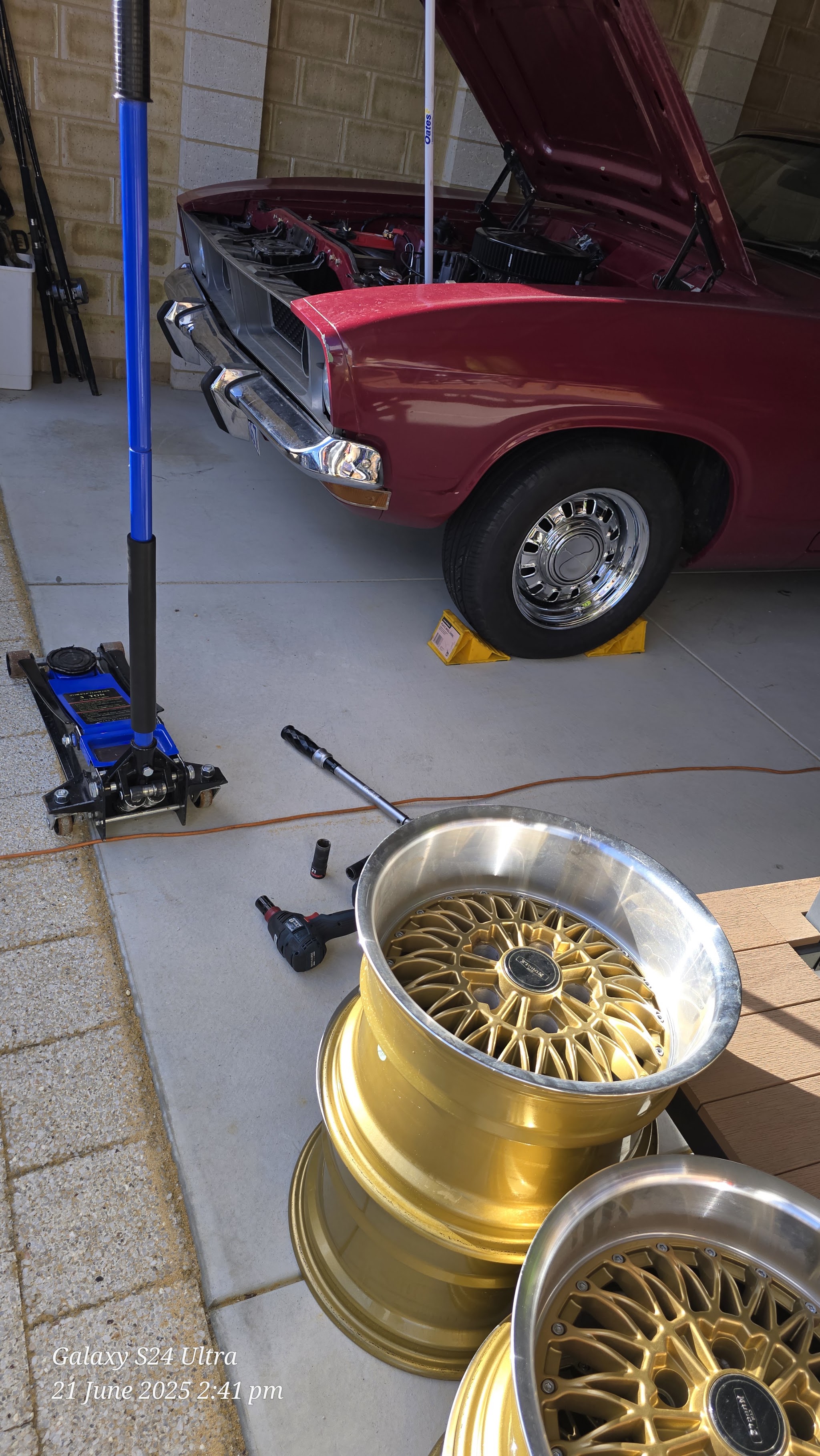 A classic maroon car on stands in a garage with a set of gold wheels ready to fit